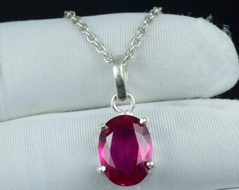 May include: A silver pendant necklace featuring a large, oval-cut ruby gemstone. The ruby is a deep, vibrant red color and is set in a four-prong silver setting. The chain is a delicate silver link. The necklace is displayed on a white surface.