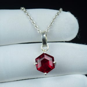 May include: A silver necklace with a hexagonal, faceted, deep red pendant. The pendant hangs from a delicate chain and has small silver accents at each point. The gemstone is a vibrant, eye-catching colour.