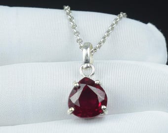 May include: A silver pendant necklace with a pear-shaped, deep red gemstone. The gemstone is set in a silver bezel and hangs from a delicate silver chain. The necklace is shown on a white surface.
