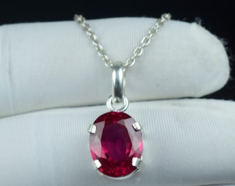 May include: A silver pendant necklace featuring an oval-cut, deep red gemstone. The gemstone is set in a silver bezel and suspended from a delicate silver chain. The background is a white fabric.
