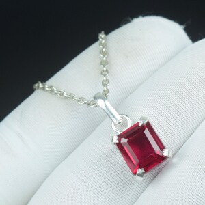 Può includere: Una collana con pendente in argento con una gemma rettangolare rosso intenso. La gemma è incastonata in una lunetta d'argento e sospesa a una delicata catena d'argento. La collana è mostrata su uno sfondo bianco.