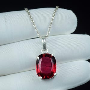 May include: A silver necklace with a large, oval-cut, deep red gemstone pendant. The gemstone is set in a silver bezel and hangs from a delicate silver chain. The background is a white surface.