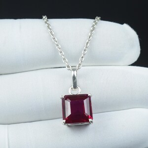 以下が含まれることがあります： 正方形の、深い赤色の宝石ペンダントが付いたシルバーネックレス。ペンダントはシルバーのフレームにセットされ、繊細なシルバーチェーンから吊り下げられています。宝石は鮮やかで光沢があり、光を反射しています。
