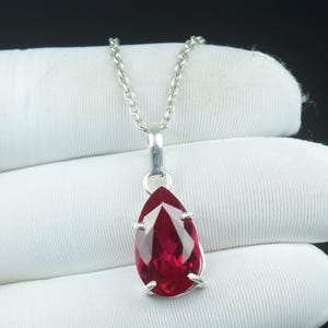 Op de afbeelding: Een zilveren hangerketting met een peervormige, gefacetteerde, dieprode edelsteen. De edelsteen wordt vastgehouden door vier zilveren tanden en hangt aan een fijne zilveren ketting. De achtergrond is een witte stof.