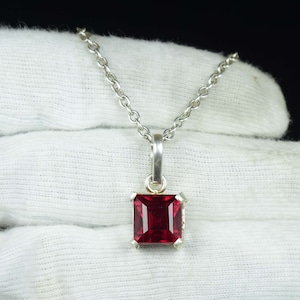 May include: A silver pendant necklace featuring a square, deep red gemstone. The gemstone is set in a square silver frame and hangs from a delicate silver chain. The necklace is displayed against a white background.