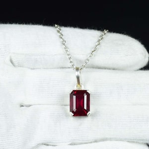 Può includere: Una collana con pendente in argento con una gemma rettangolare rosso intenso. La gemma è incastonata in una cornice d'argento e appesa a una delicata catena d'argento. La collana è esposta su una superficie bianca.