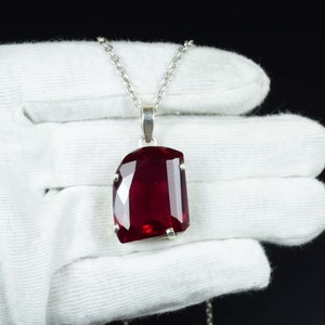 Puede incluir: Un collar de plata con un colgante grande, rectangular, facetado y de color rojo intenso. El colgante está sujeto por cuatro garras de plata. El collar se muestra sobre un fondo negro.