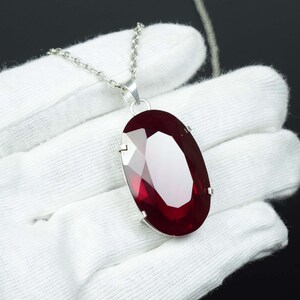 May include: A silver necklace featuring a large, oval-cut, deep red gemstone pendant. The faceted gemstone reflects light, creating a brilliant shine. The chain is fine, and the pendant is set in a silver bezel.