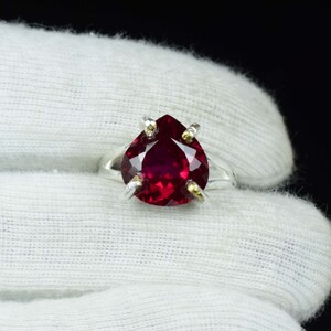 May include: A silver ring featuring a large, pear-shaped, deep red gemstone. The gemstone is held in place by four prongs with gold accents. The ring is set against a white background.