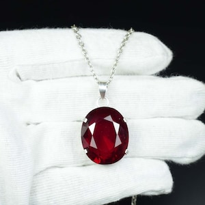 May include: A silver necklace with a large, oval-cut, deep red gemstone pendant. The faceted gemstone reflects light, creating a brilliant shine. The chain is fine, and the pendant is the main feature of the jewellery.