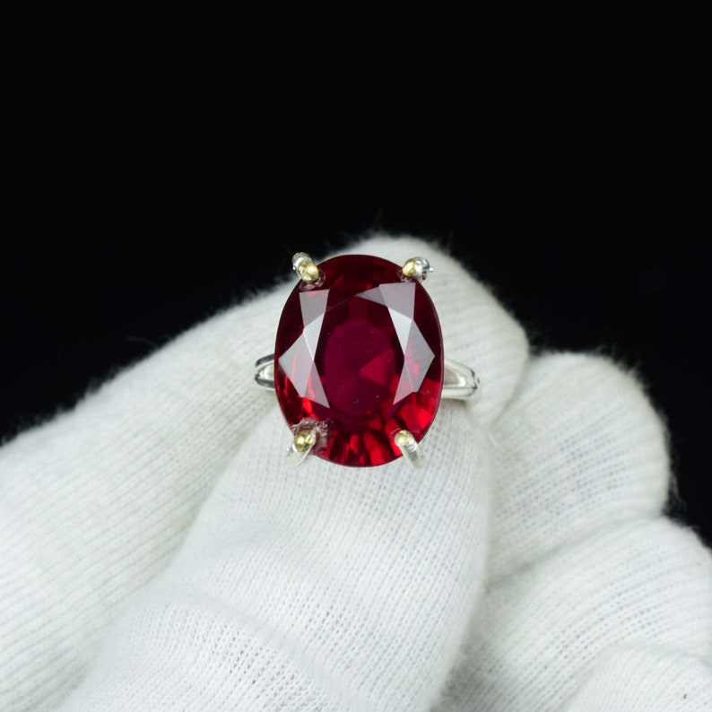 Pode incluir: Um anel de prata com uma grande pedra preciosa vermelha escura de corte oval. A pedra &eacute; presa por quatro garras com detalhes dourados. O anel est&aacute; sobre um fundo branco com um fundo preto.