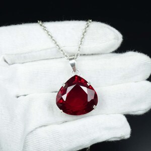 May include: A silver necklace featuring a large, teardrop-shaped, faceted, deep red gemstone pendant. The pendant is set in a silver bezel and hangs from a delicate silver chain. The background is black.