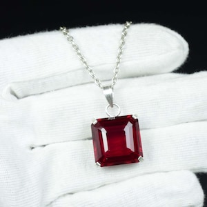 Op de afbeelding: Een zilveren ketting met een rechthoekige, dieprode edelsteen hanger. De hanger is gezet in een zilveren montuur en hangt aan een fijne zilveren ketting. De edelsteen heeft een facetgeslepen oppervlak. De sieraden worden tegen een witte achtergrond getoond.