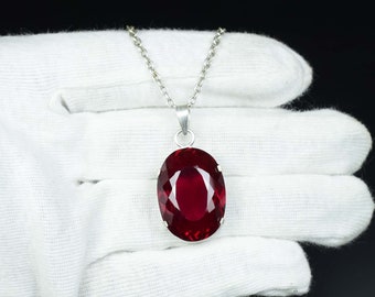May include: A silver pendant necklace featuring a large, oval-cut, deep red gemstone. The gemstone is set in a silver bezel and hangs from a delicate silver chain. The background is a white fabric.