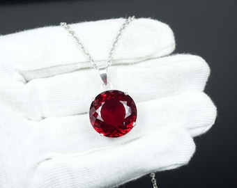 Puede incluir: Un collar de plata con un colgante de piedra preciosa roja oscura, grande, redondo y facetado. El colgante está engastado en un bisel de plata y cuelga de una delicada cadena de plata. La piedra preciosa tiene un corte brillante.