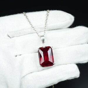 May include: A silver necklace featuring a large, rectangular, deep red gemstone pendant. The pendant is set in a silver frame and hangs from a delicate silver chain. The gemstone has a faceted cut, reflecting light. The background is a white glove.