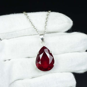 May include: A silver necklace featuring a large, faceted, teardrop-shaped, deep red pendant. The pendant is set in a silver bezel and hangs from a delicate silver chain. The background is a black surface and a white glove.