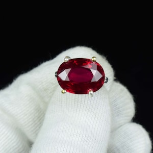 May include: A close-up of a ring featuring a large, oval-cut ruby gemstone. The ruby is a deep red colour and is set in a silver band with gold accents. The ring is held against a white background.