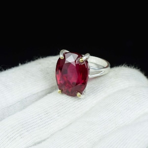 Pode incluir: Um anel de prata com uma grande pedra preciosa vermelha escura de corte oval. A pedra &eacute; mantida no lugar por quatro garras com detalhes dourados. O anel est&aacute; sobre um fundo de tecido branco com um fundo preto.