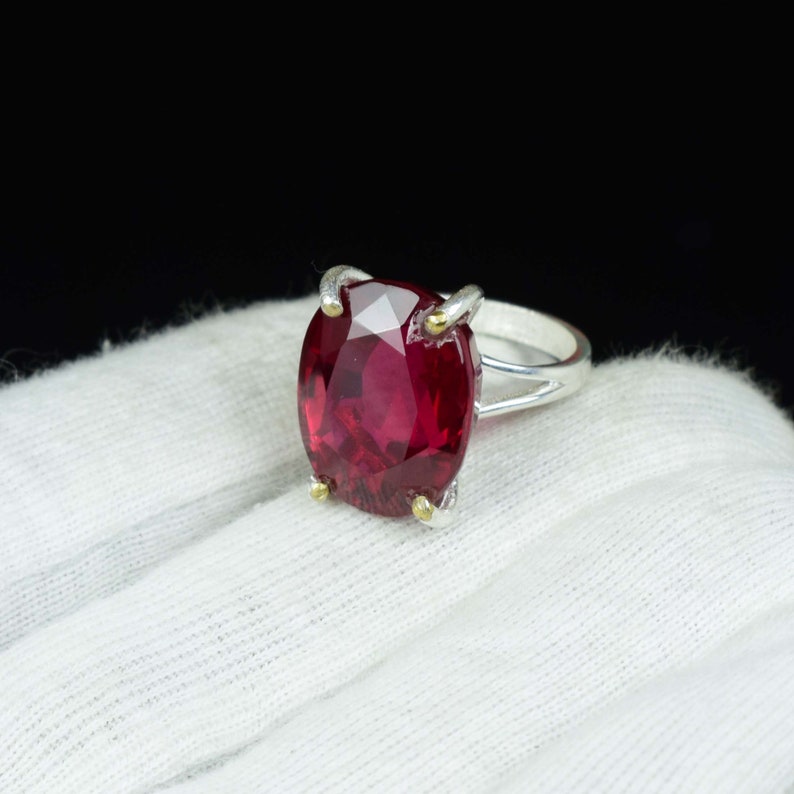 Pode incluir: Um anel de prata com uma grande pedra preciosa vermelha escura de corte oval. A pedra &eacute; mantida no lugar por quatro garras com detalhes dourados. O anel est&aacute; sobre um fundo de tecido branco com um fundo preto.