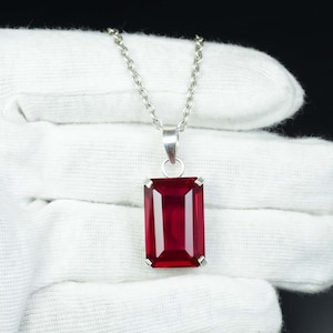 Pode incluir: Colar com pingente retangular vermelho escuro e corrente prateada. O pingente é uma grande pedra preciosa facetada emoldurada em uma moldura prateada. O colar é exibido em um fundo branco.