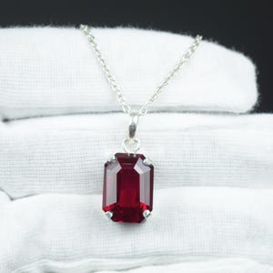 Op de afbeelding: Een zilveren ketting met een rechthoekige, dieprode hanger. De hanger is een gefacetteerde edelsteen, gezet in een zilveren montuur. De ketting is delicaat en de hanger is het middelpunt van de sieraden.