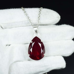 May include: A silver necklace with a large, teardrop-shaped, faceted, deep red pendant. The pendant is held by a silver bail and hangs from a delicate silver chain. The necklace is displayed against a white background.