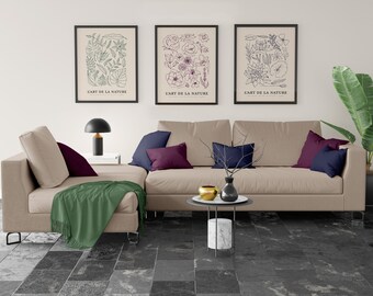 May include: A beige sectional sofa with three throw pillows, two dark purple and one navy blue, and a green throw blanket. The sofa is in front of a white wall with three framed black and white botanical prints with the text "L'ART DE LA NATURE".