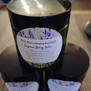 May include: Three dark green jars of Rosa Mae's Healing Essentials Eczema Relief Salve. Each jar has a black lid and a white label with text and a lavender design. The label lists ingredients including plantain, echinacea, and rosemary. Each jar contains 4 oz.