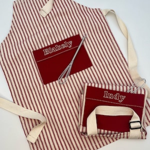 May include: Red and white striped apron with a red pocket embroidered with the name "Blakely". A smaller matching apron with the name "Indy" is also visible. Both aprons have cream-colored straps and a small whisk.