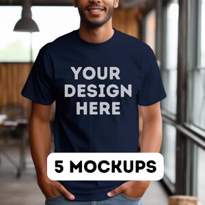 Può includere: Una t-shirt blu navy con il testo "YOUR DESIGN HERE" stampato in bianco sul davanti. La t-shirt è indossata da una persona in piedi in una stanza con una finestra sullo sfondo. Il testo "5 MOCKUPS" è stampato in bianco su un rettangolo nero nella parte inferiore dell'immagine.