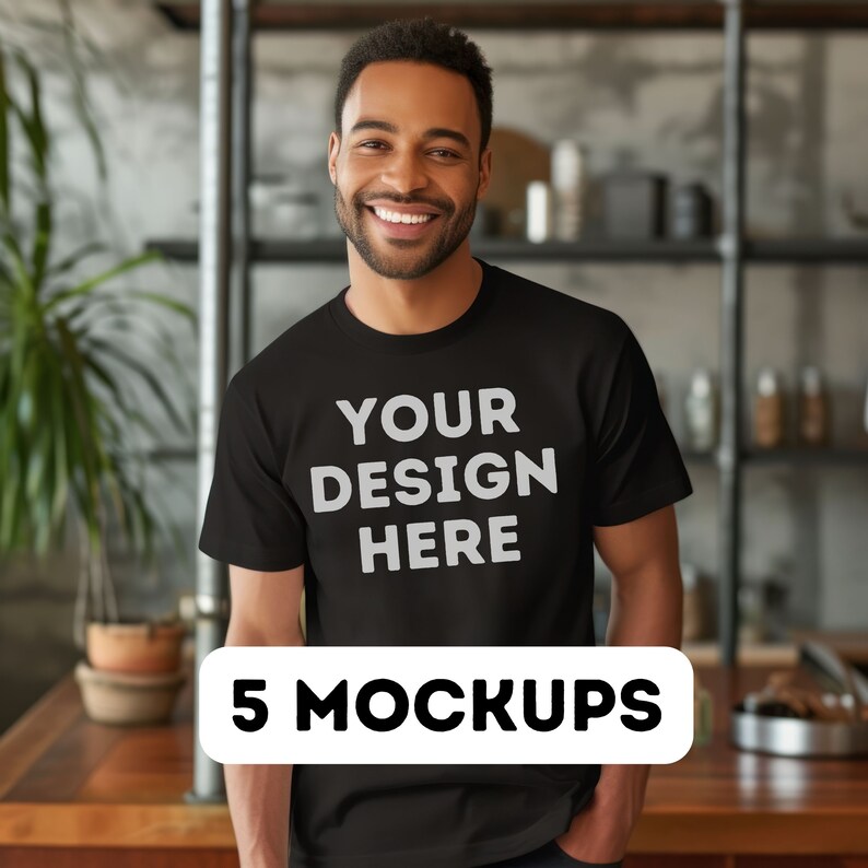 以下が含まれることがあります： 「YOUR DESIGN HERE」という文字が書かれた黒いTシャツのモックアップ。Tシャツは笑顔の人物が着用しています。背景はぼやけたキッチンです。