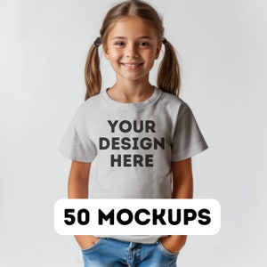 May include: A young girl wearing a light gray t-shirt with the text "YOUR DESIGN HERE" in black. Below the text is a white rectangle with the words "50 MOCKUPS" in black. The girl is smiling and wearing blue jeans.