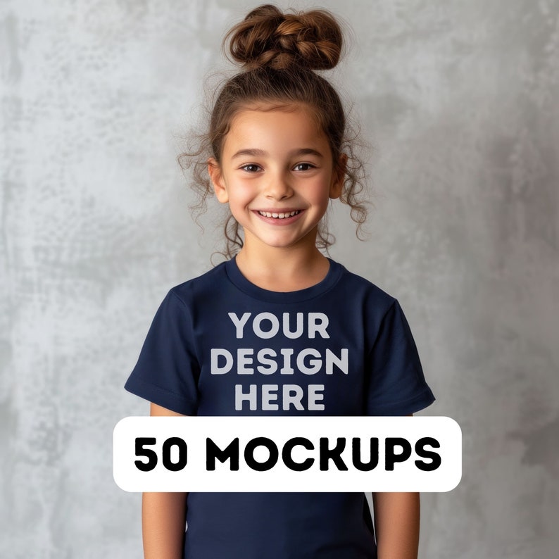 May include: A young girl smiles while wearing a navy blue t-shirt. The shirt has the text "YOUR DESIGN HERE" printed on the front, with a white banner below that reads "50 MOCKUPS". The background is a mottled gray.