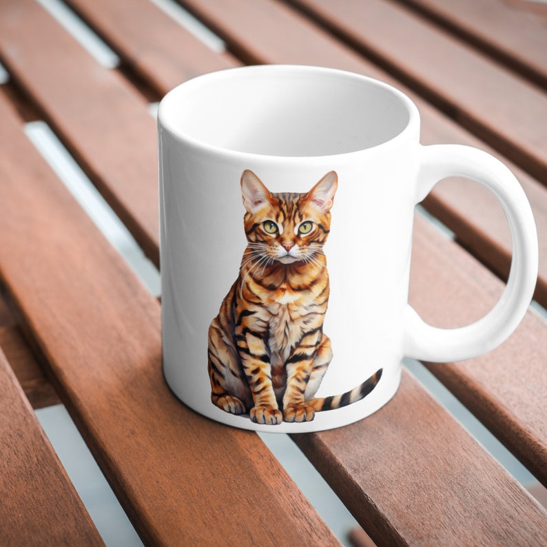 May include: White ceramic mug with a brown and black tabby cat sitting on it. The cat is looking directly at the camera.