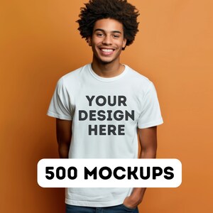 以下が含まれることがあります： 白いTシャツに「YOUR DESIGN HERE」という文字が前面に印刷されています。Tシャツは、オレンジ色の背景に、縮れた髪の毛の人によって着用されています。500 MOCKUPS.