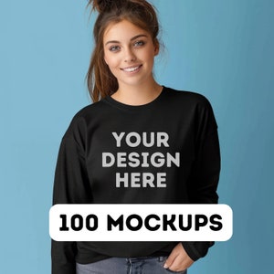 May include: A black sweatshirt with the text "YOUR DESIGN HERE" printed on the front. The text is in a light gray color. The sweatshirt is being modeled by a person with long brown hair. The background is a light blue color. 100 MOCKUPS.