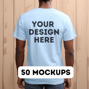 May include: A light blue t-shirt with the text "YOUR DESIGN HERE" printed on the back. The t-shirt is being worn by a person standing in front of a wood panel background. 50 MOCKUPS.