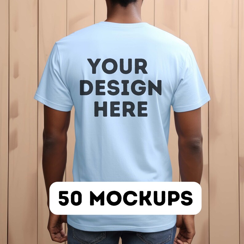 May include: A light blue t-shirt with the text "YOUR DESIGN HERE" printed on the back. The t-shirt is being worn by a person standing in front of a wood panel background. 50 MOCKUPS.
