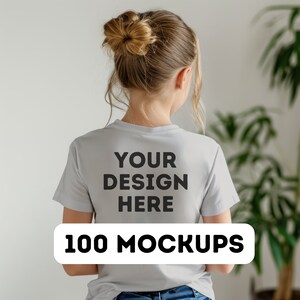 Pu&ograve; includere: Una maglietta grigio chiaro con la scritta "YOUR DESIGN HERE" stampata sul retro in lettere nere. Sotto, un rettangolo bianco con le parole "100 MOCKUPS" in nero. La persona &egrave; di spalle.