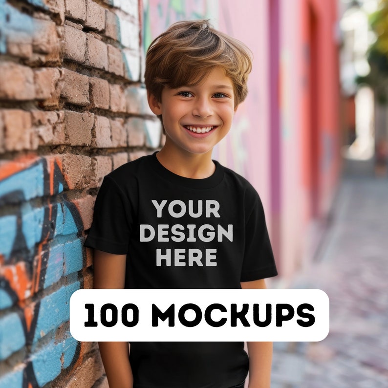 Pu&ograve; includere: Un giovane indossa una maglietta nera con la scritta bianca "YOUR DESIGN HERE". Sotto, un banner bianco recita "100 MOCKUPS". La persona sorride, appoggiata a un muro di mattoni con graffiti. Lo sfondo &egrave; sfocato.