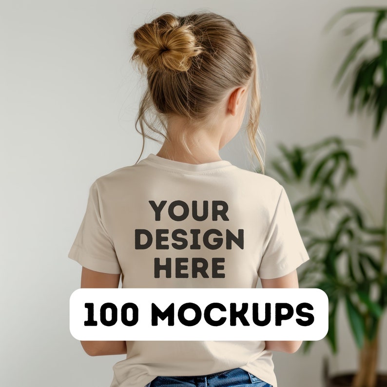 May include: A beige t-shirt with the text "YOUR DESIGN HERE" printed on the back in black, with a white rectangle below that reads "100 MOCKUPS". The t-shirt is being worn by a person with their hair up in a bun.