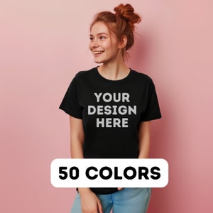 以下が含まれることがあります： 前面に「YOUR DESIGN HERE」という文字が印刷された黒いTシャツ。赤い髪の女性が着用しています。背景はピンクです。 50色