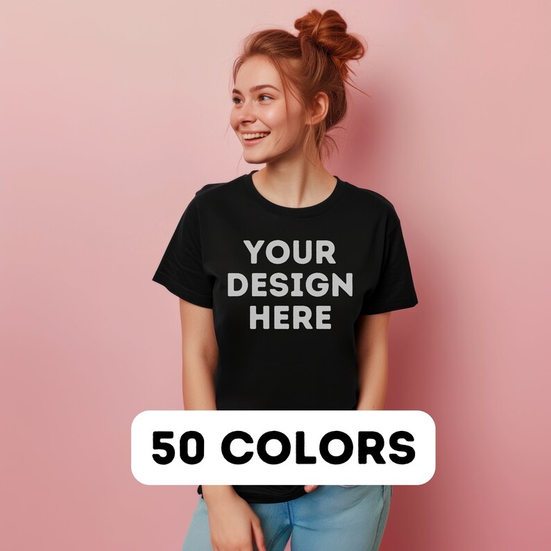 以下が含まれることがあります： 前面に「YOUR DESIGN HERE」という文字が印刷された黒いTシャツ。赤い髪の女性が着用しています。背景はピンクです。 50色