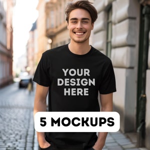 以下が含まれることがあります： 前面に「YOUR DESIGN HERE」という文字が印刷された黒いTシャツ。Tシャツは街の通りの真ん中に立っている人が着ています。5 MOCKUPS.