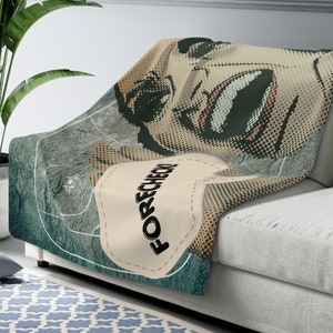 May include: A green and beige throw blanket with a pop art style illustration of a woman's face and the word "FORECHECK" in a heart shape.