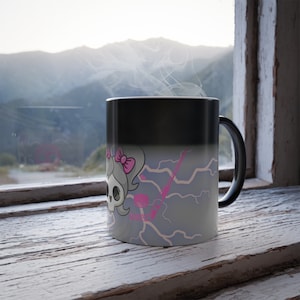 May include: A black ceramic mug that transitions to a lighter gray color, featuring a graphic of a skull with a pink bow, a pink hockey stick, and a pink puck, surrounded by light pink lightning bolt patterns against a gray background.