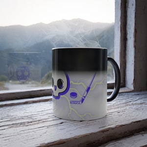 May include: A black ceramic mug with a heat-sensitive surface that reveals a white graphic featuring a purple skull, a hockey stick, a hockey puck, and yellow lightning bolts. The mug has a curved handle and a smooth, cylindrical body design.