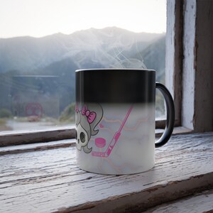 May include: A black ceramic mug with a heat-sensitive surface that reveals a graphic of a skull with a pink bow, a pink hockey stick, and a pink puck against a light background with faint orange lightning bolt patterns and steam rising.