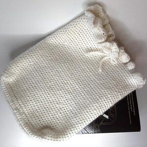 May include: A white crocheted drawstring pouch featuring a textured stitch pattern and a ruffled top edge. The pouch is partially covering a black book with visible white text that reads, "full of unique magic" and "bestselling author of the inheritance cycle."